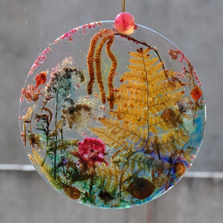 Transparentny roślinny medalion o numerze 13 zawieszony w oknie. Światło słoneczne podświetla rośliny zatopione w żywicy.