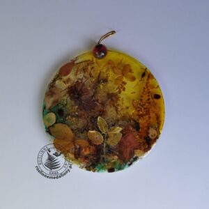 Ręcznie wykonany, okrągły medalion z żywicy, w którym zatopiono różnobarwne suszone rośliny, umieszczony na kartce papieru.