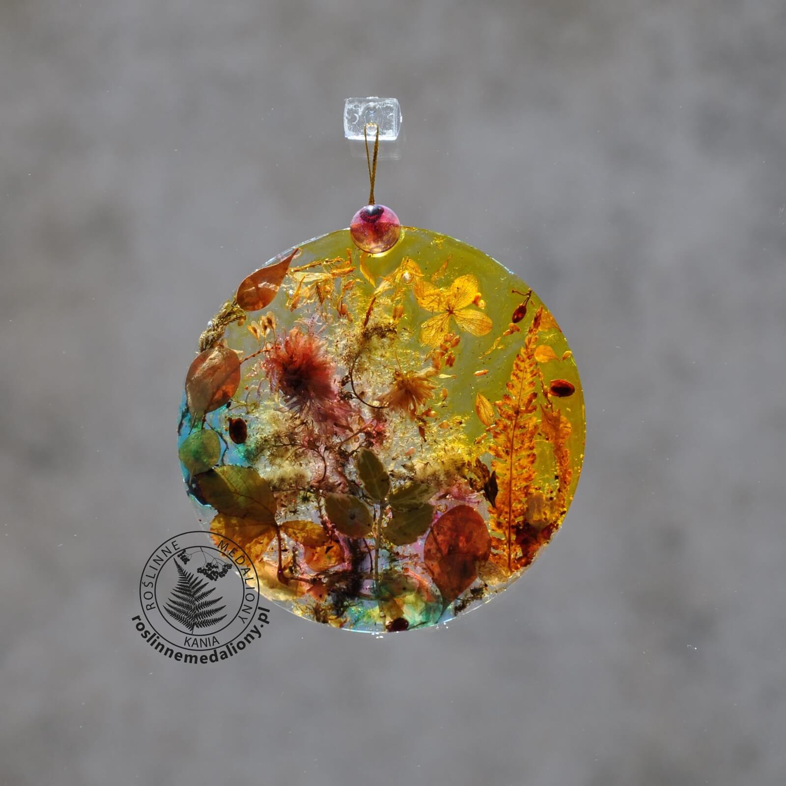 Ręcznie wykonany, okrągły medalion z żywicy, w którym zatopiono różnobarwne suszone rośliny, zawieszony na szybie okna.