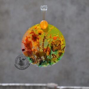 Ręcznie wykonany, okrągły medalion z żywicy, w którym zatopiono różnobarwne suszone rośliny, zawieszony na szybie okna.