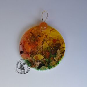 Ręcznie wykonany, okrągły medalion z żywicy, w którym zatopiono różnobarwne suszone rośliny, umieszczony na kartce papieru.