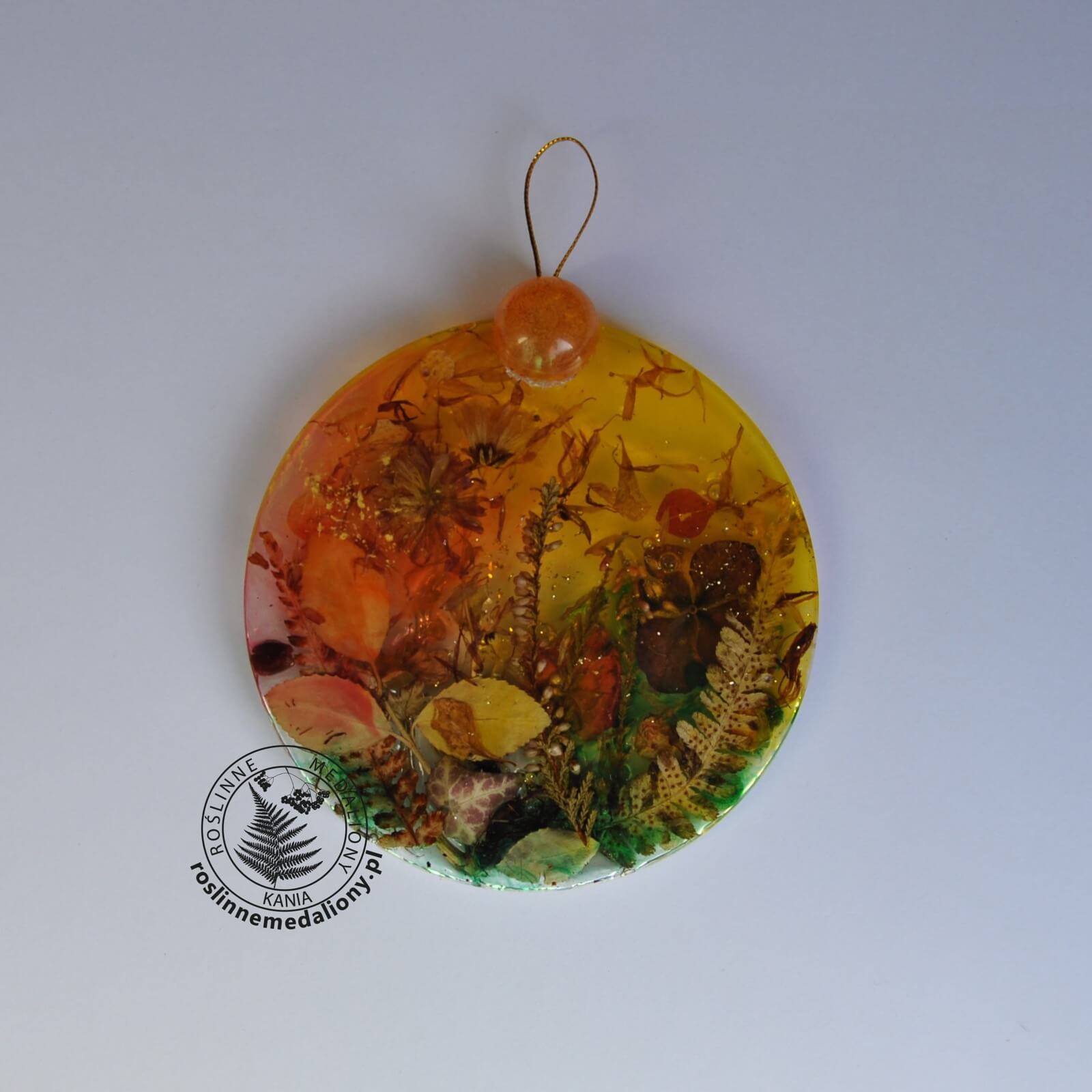 Ręcznie wykonany, okrągły medalion z żywicy, w którym zatopiono różnobarwne suszone rośliny, umieszczony na kartce papieru.