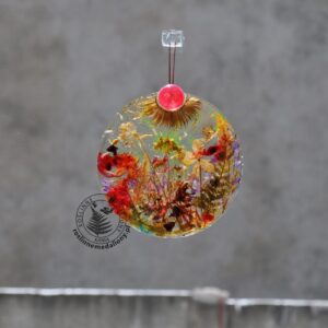 Ręcznie wykonany, okrągły medalion z żywicy, w którym zatopiono różnobarwne suszone rośliny, zawieszony na szybie okna.