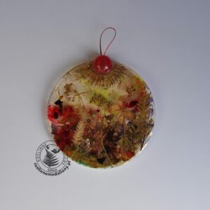 Ręcznie wykonany, okrągły medalion z żywicy, w którym zatopiono różnobarwne suszone rośliny, umieszczony na kartce papieru.