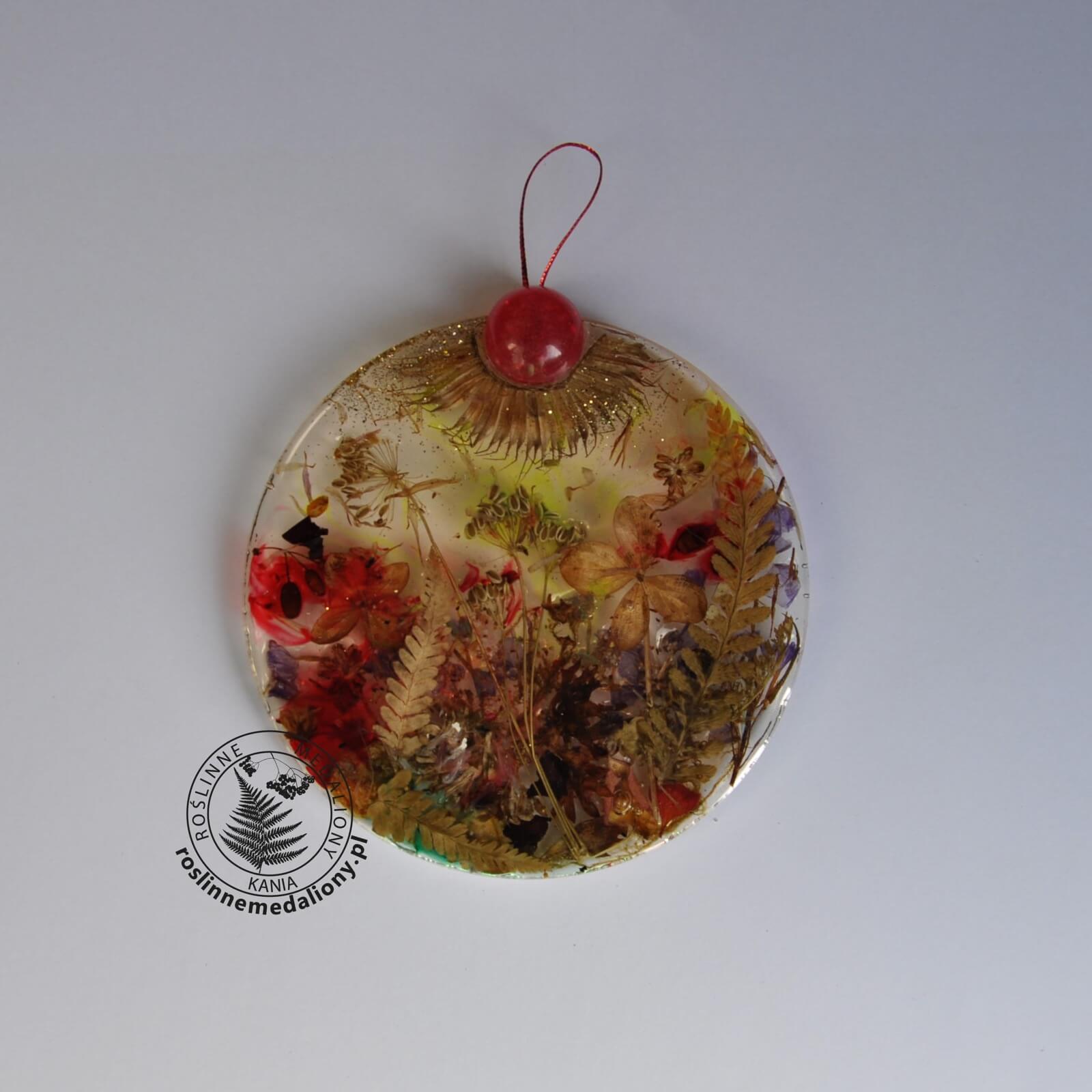 Ręcznie wykonany, okrągły medalion z żywicy, w którym zatopiono różnobarwne suszone rośliny, umieszczony na kartce papieru.