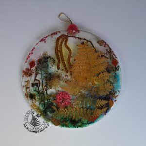 Ręcznie wykonany, okrągły medalion z żywicy, w którym zatopiono różnobarwne suszone rośliny, umieszczony na kartce papieru.