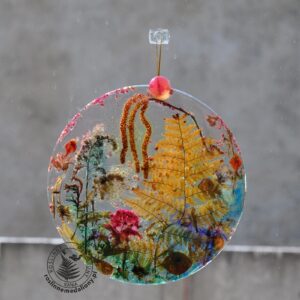 Ręcznie wykonany, okrągły medalion z żywicy, w którym zatopiono różnobarwne suszone rośliny, zawieszony na szybie okna.