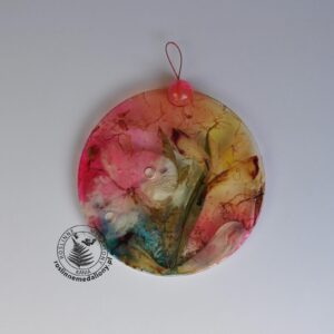 Ręcznie wykonany, okrągły medalion z żywicy, w którym zatopiono różnobarwne suszone rośliny, umieszczony na kartce papieru.