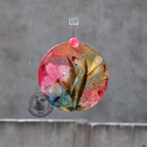 Ręcznie wykonany, okrągły medalion z żywicy, w którym zatopiono różnobarwne suszone rośliny, zawieszony na szybie okna.