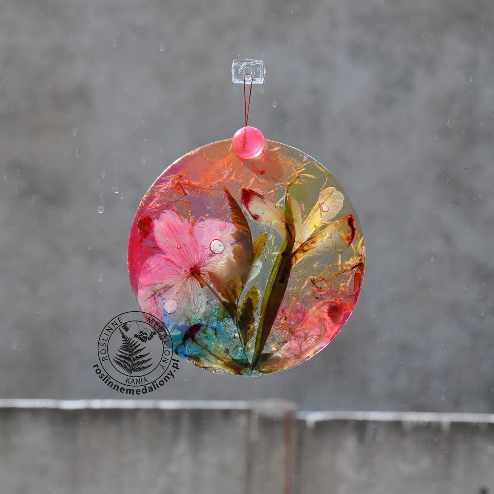 Ręcznie wykonany, okrągły medalion z żywicy, w którym zatopiono różnobarwne suszone rośliny, zawieszony na szybie okna.