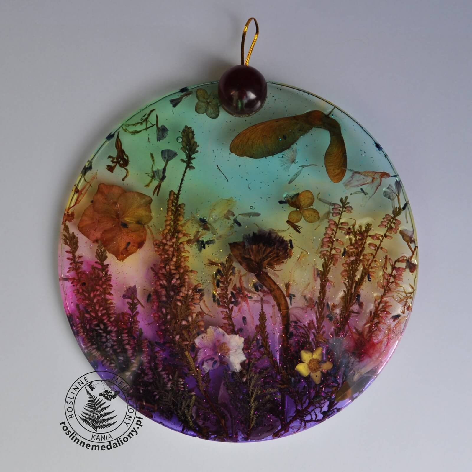 Ręcznie wykonany, okrągły medalion z żywicy, w którym zatopiono różnobarwne suszone rośliny, umieszczony na kartce papieru.