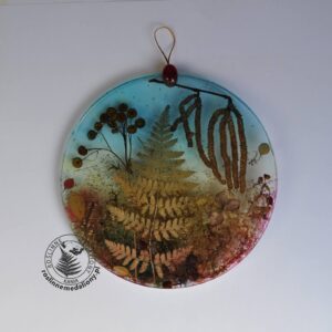 Ręcznie wykonany, okrągły medalion z żywicy, w którym zatopiono różnobarwne suszone rośliny, umieszczony na kartce papieru.