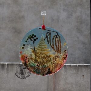 Ręcznie wykonany, okrągły medalion z żywicy, w którym zatopiono różnobarwne suszone rośliny, w tym liście paproci i bratki, zawieszony na szybie okna.