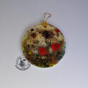 Ręcznie wykonany, okrągły medalion z żywicy, w którym zatopiono różnobarwne suszone rośliny, umieszczony na kartce papieru.