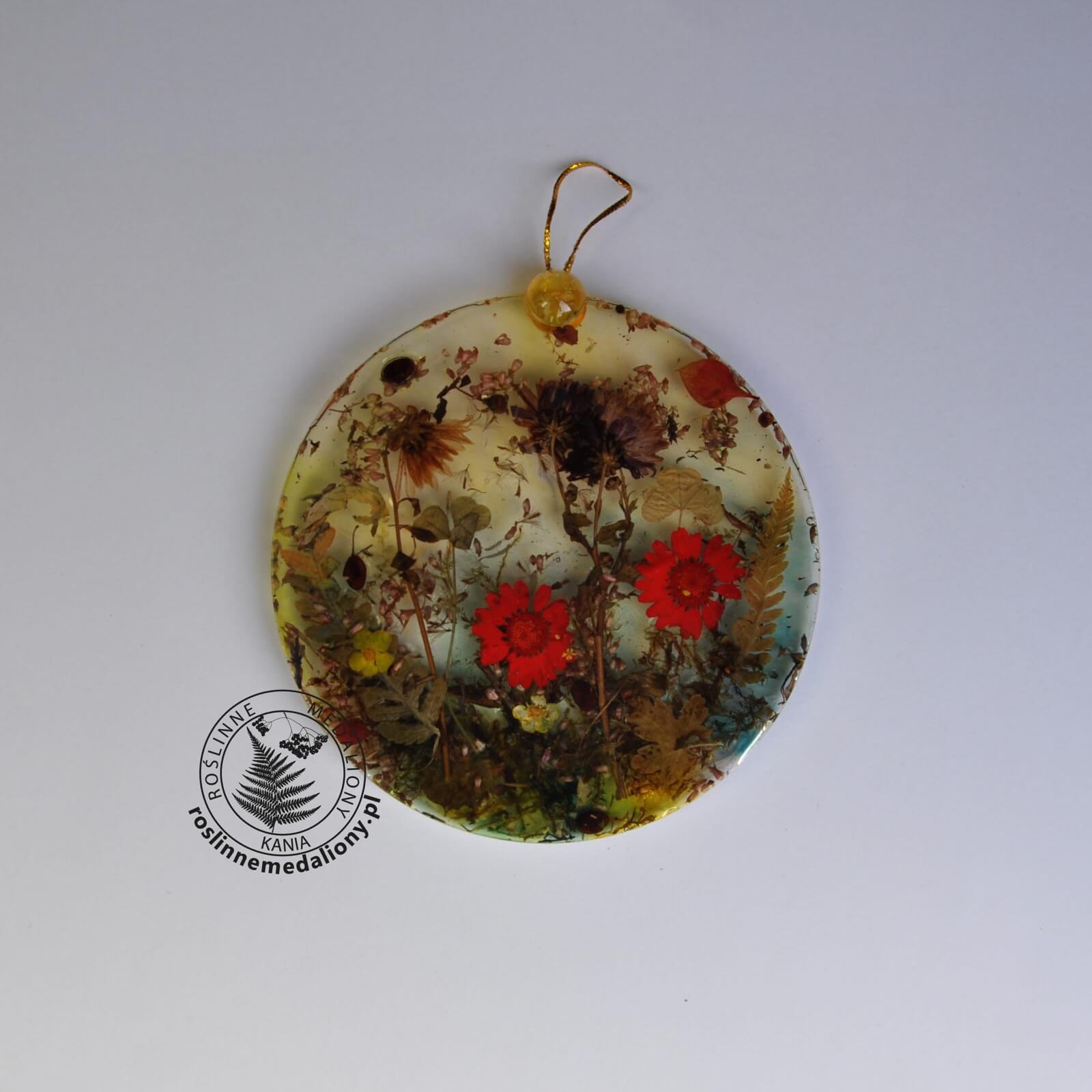 Ręcznie wykonany, okrągły medalion z żywicy, w którym zatopiono różnobarwne suszone rośliny, umieszczony na kartce papieru.