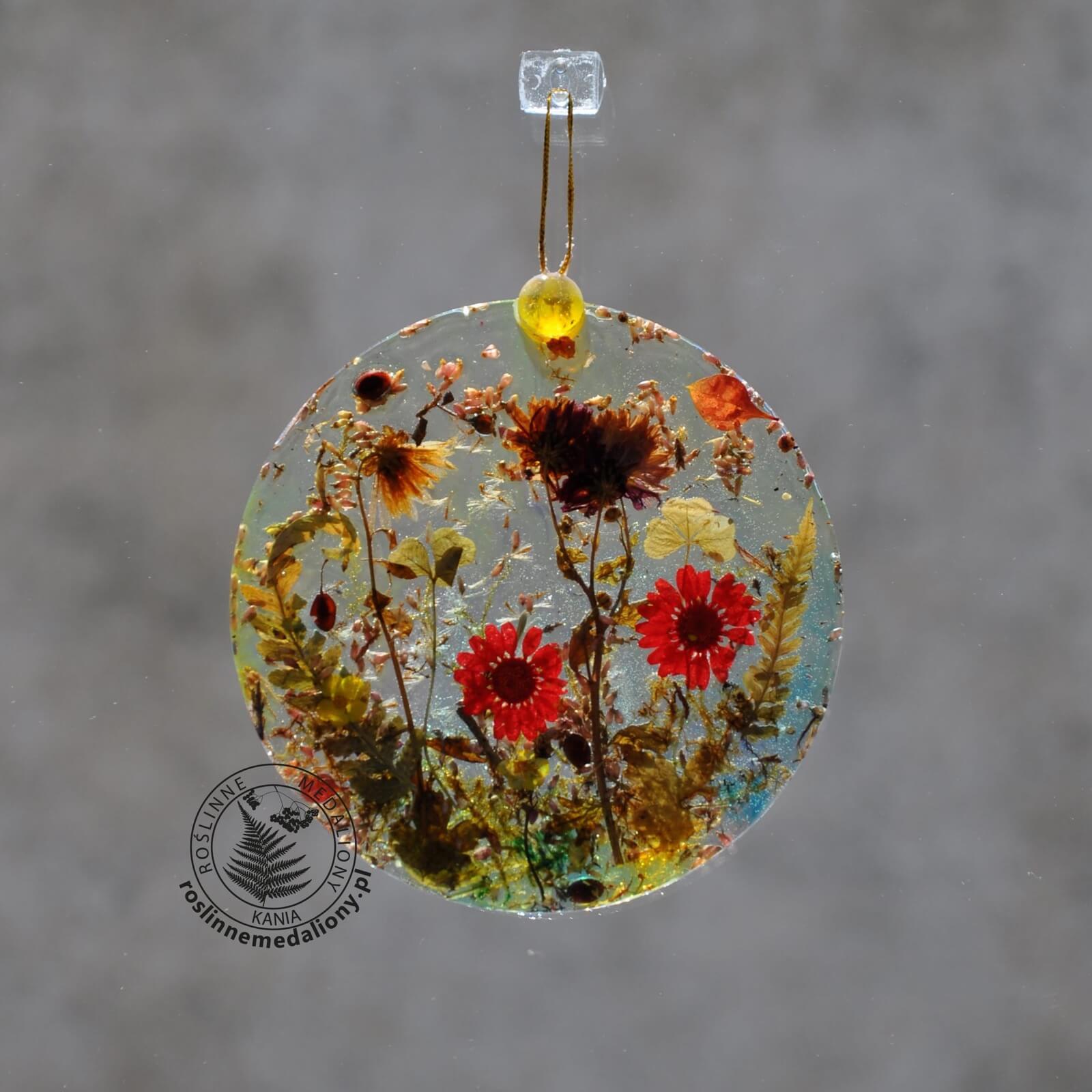Ręcznie wykonany, okrągły medalion z żywicy, w którym zatopiono różnobarwne suszone rośliny, w tym liście paproci i bratki, zawieszony na szybie okna.