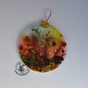 Ręcznie wykonany, okrągły medalion z żywicy, w którym zatopiono różnobarwne suszone rośliny, umieszczony na kartce papieru.