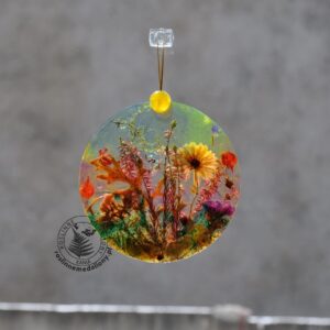 Ręcznie wykonany, okrągły medalion z żywicy, w którym zatopiono różnobarwne suszone rośliny, zawieszony na szybie okna.