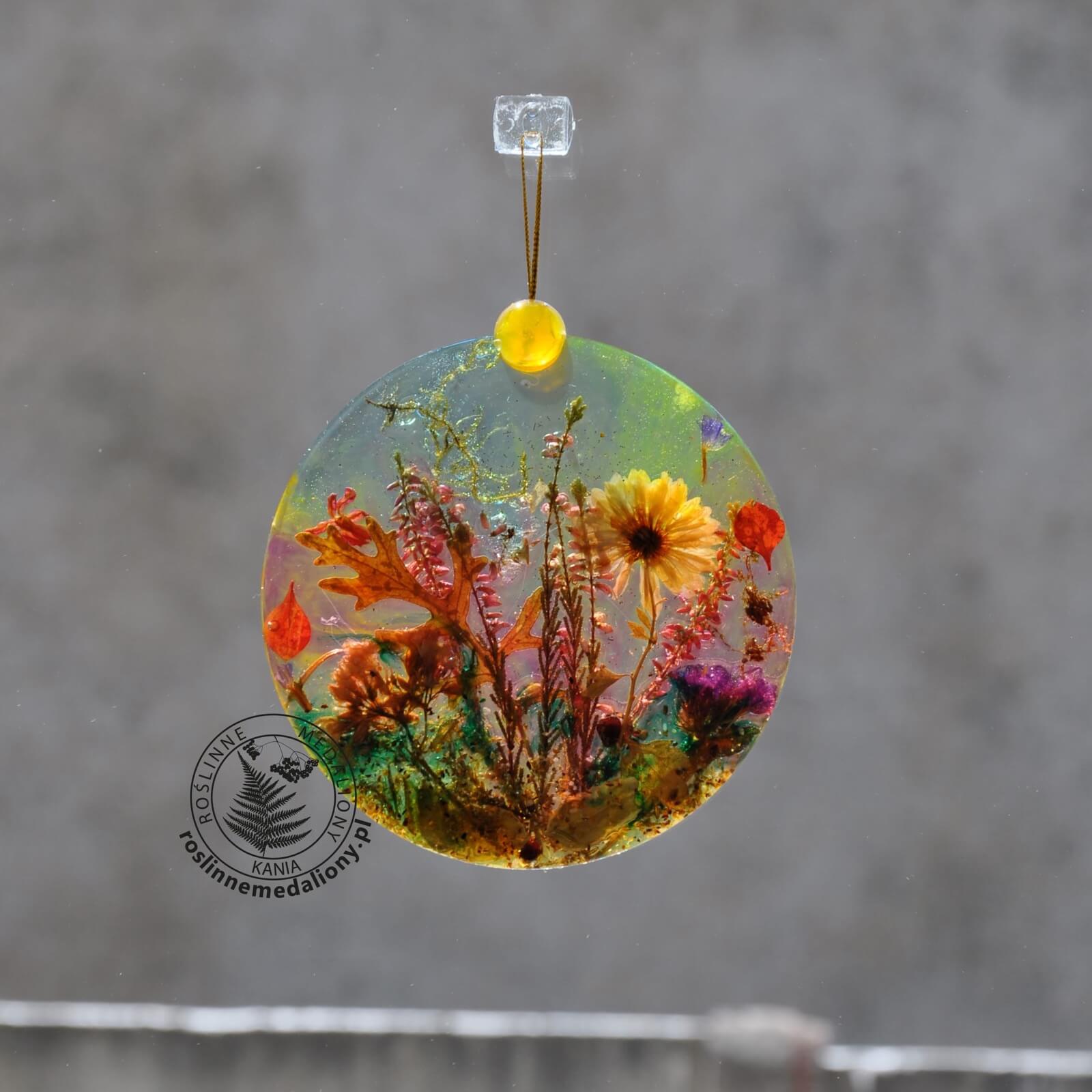 Ręcznie wykonany, okrągły medalion z żywicy, w którym zatopiono różnobarwne suszone rośliny, zawieszony na szybie okna.