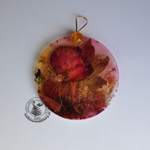 Ręcznie wykonany, okrągły medalion z żywicy, w którym zatopiono różnobarwne suszone rośliny, umieszczony na kartce papieru.