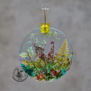 Ręcznie wykonany, okrągły medalion z żywicy, w którym zatopiono różnobarwne suszone rośliny, zawieszony na szybie okna.
