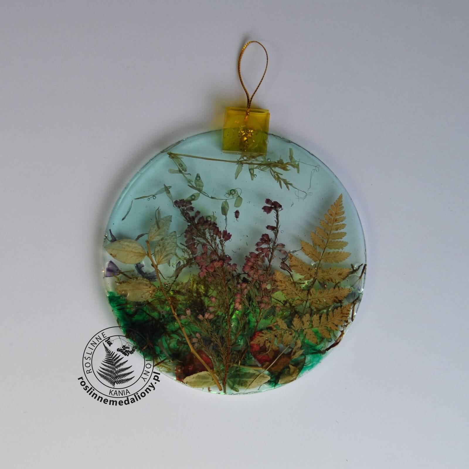Ręcznie wykonany, okrągły medalion z żywicy, w którym zatopiono różnobarwne suszone rośliny, umieszczony na kartce papieru.