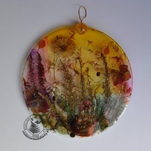 Ręcznie wykonany, okrągły medalion z żywicy, w którym zatopiono różnobarwne suszone rośliny, umieszczony na kartce papieru.