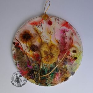 Ręcznie wykonany, okrągły medalion z żywicy, w którym zatopiono różnobarwne suszone rośliny, umieszczony na kartce papieru.