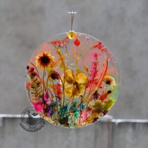 Ręcznie wykonany, okrągły medalion z żywicy, w którym zatopiono różnobarwne suszone rośliny, zawieszony na szybie okna.