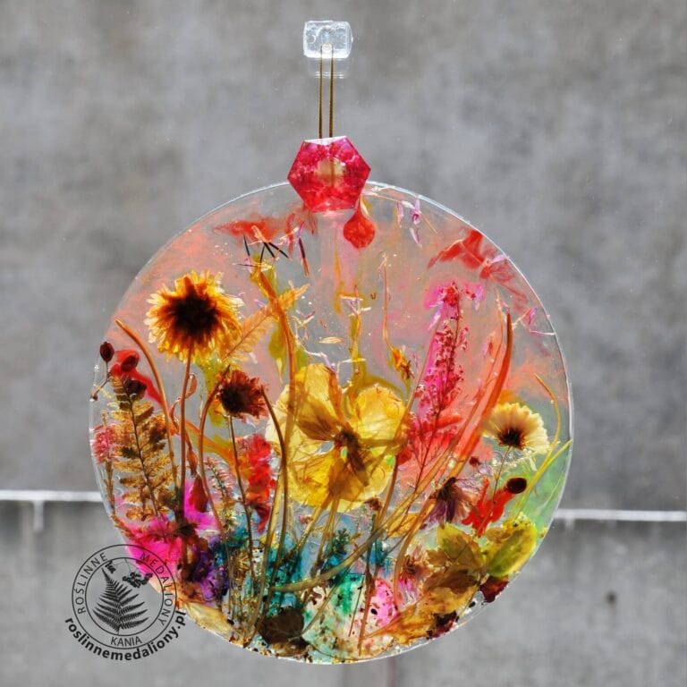 Ręcznie wykonany, okrągły medalion z żywicy, w którym zatopiono różnobarwne suszone rośliny, zawieszony na szybie okna.