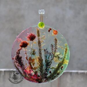 Ręcznie wykonany, okrągły medalion z żywicy, w którym zatopiono różnobarwne suszone rośliny, zawieszony na szybie okna.