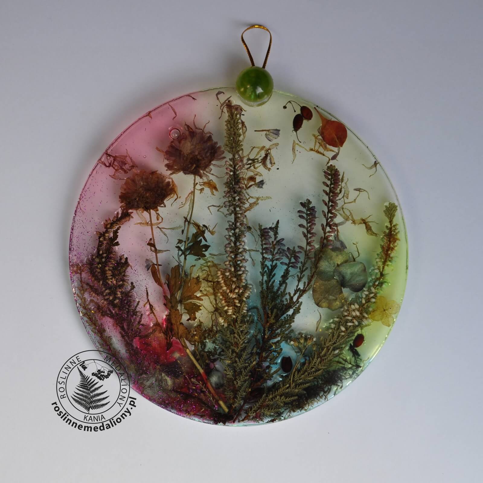 Ręcznie wykonany, okrągły medalion z żywicy, w którym zatopiono różnobarwne suszone rośliny, umieszczony na kartce papieru.