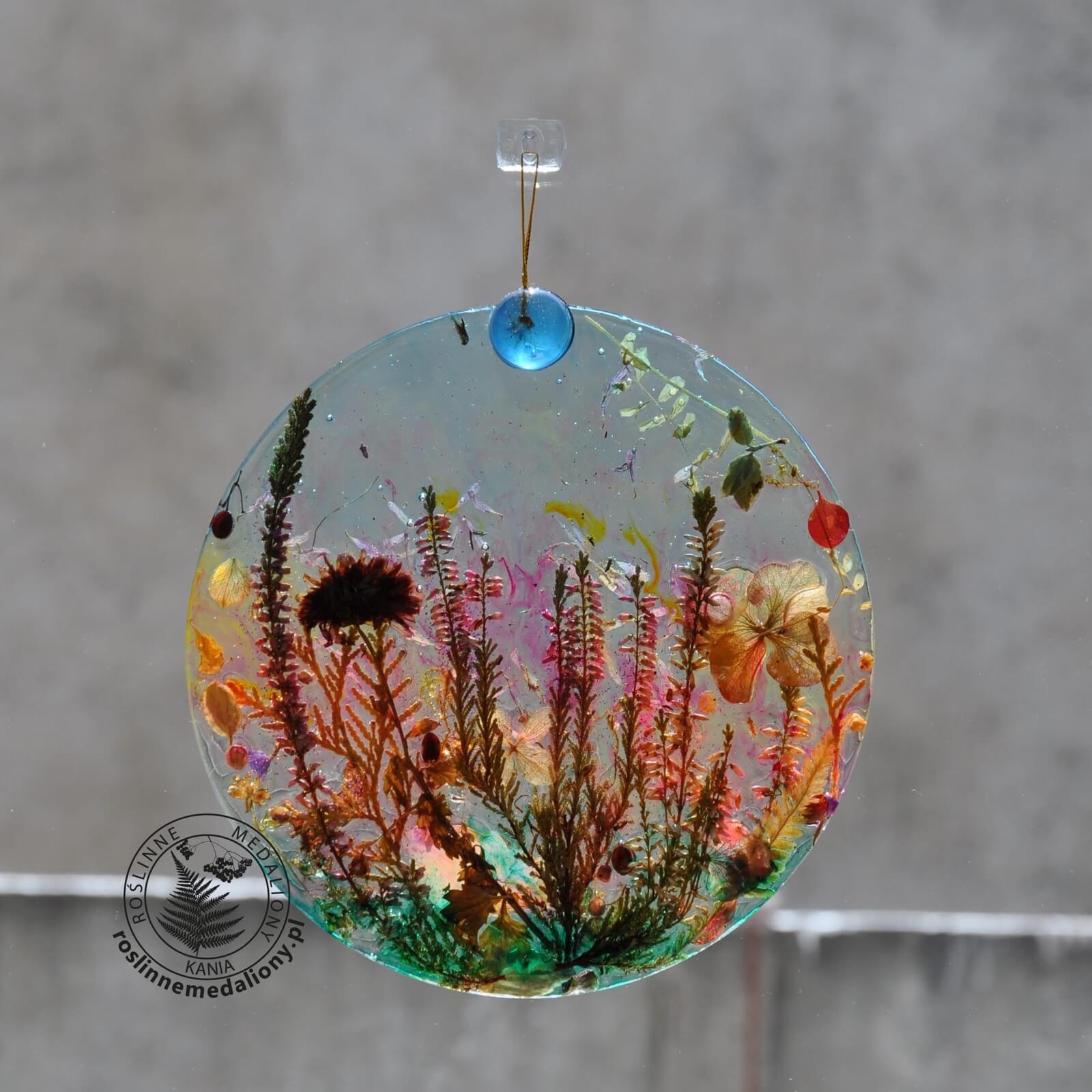 Ręcznie wykonany, okrągły medalion z żywicy, w którym zatopiono różnobarwne suszone rośliny, zawieszony na szybie okna.