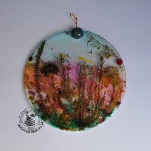 Ręcznie wykonany, okrągły medalion z żywicy, w którym zatopiono różnobarwne suszone rośliny, umieszczony na kartce papieru.