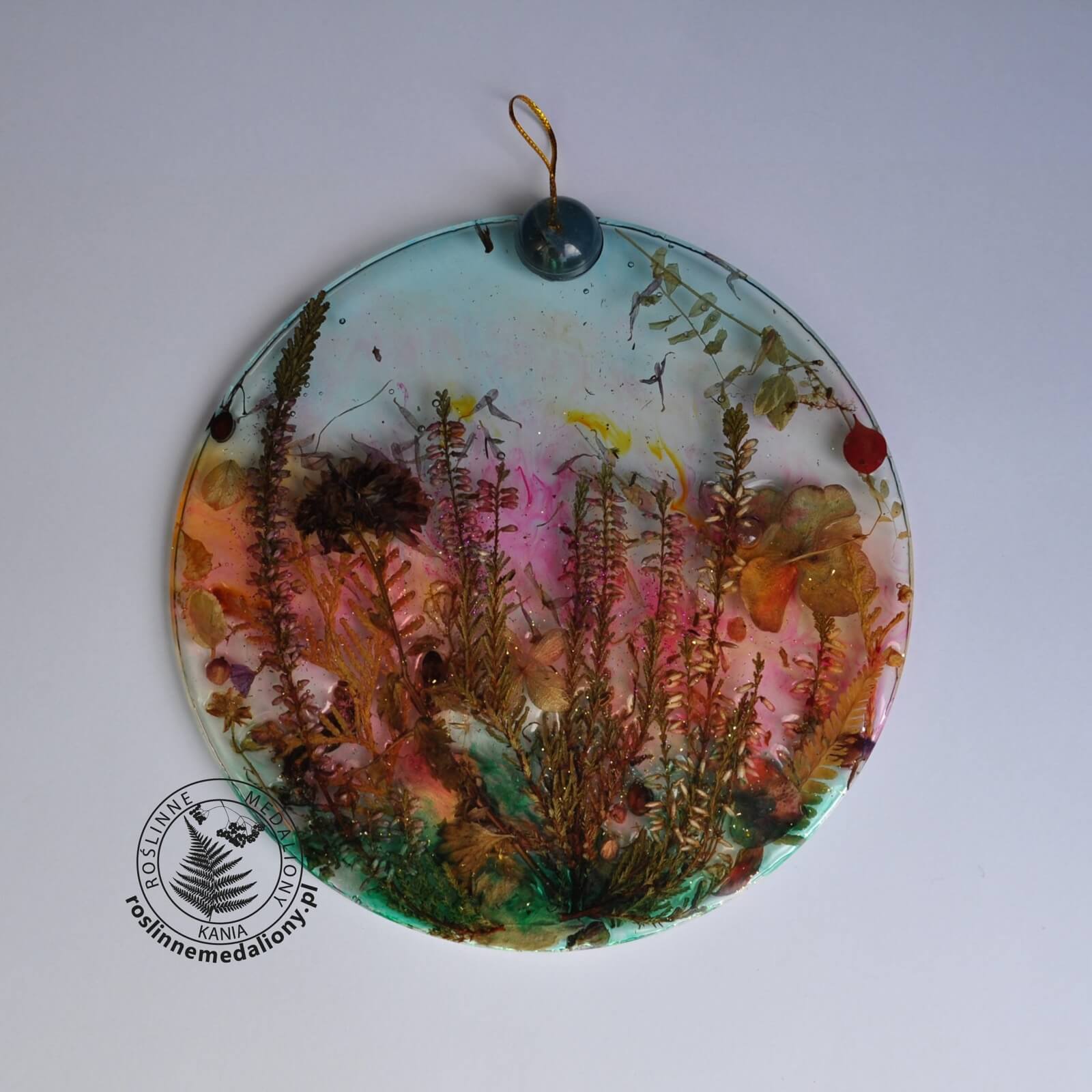 Ręcznie wykonany, okrągły medalion z żywicy, w którym zatopiono różnobarwne suszone rośliny, umieszczony na kartce papieru.