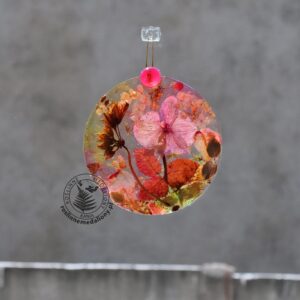 Ręcznie wykonany, okrągły medalion z żywicy, w którym zatopiono różnobarwne suszone rośliny, w tym liście paproci i bratki, zawieszony na szybie okna.