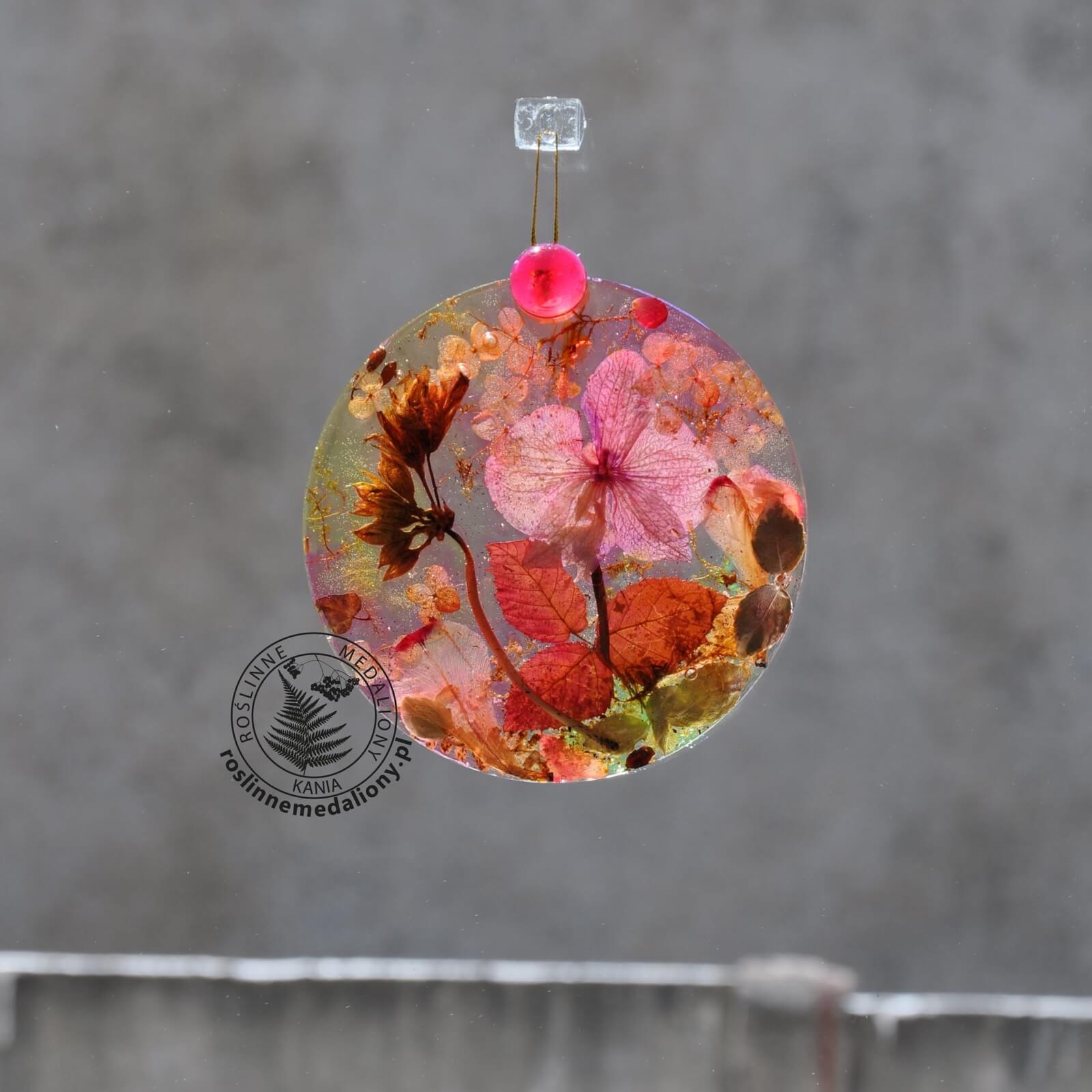 Ręcznie wykonany, okrągły medalion z żywicy, w którym zatopiono różnobarwne suszone rośliny, w tym liście paproci i bratki, zawieszony na szybie okna.