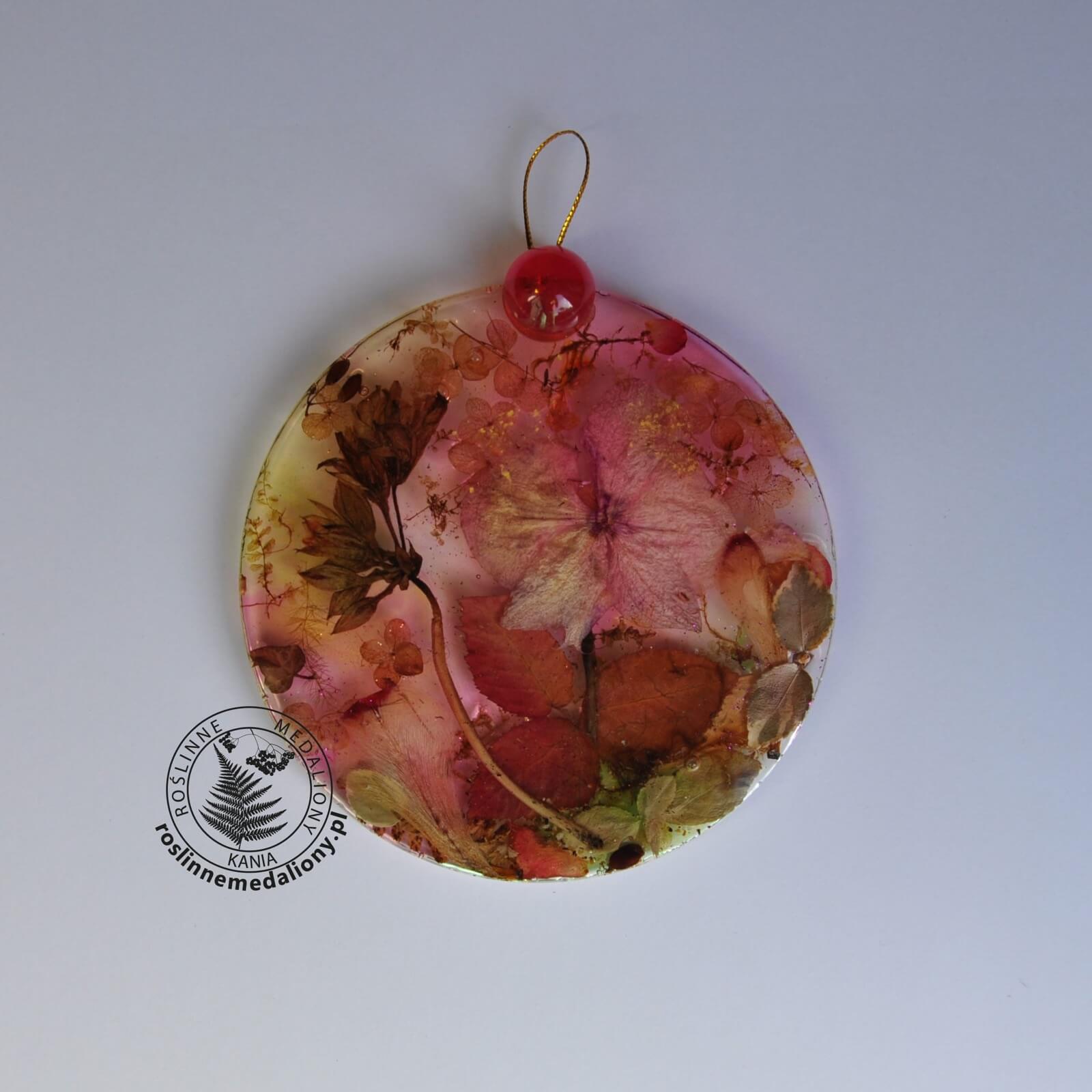 Ręcznie wykonany, okrągły medalion z żywicy, w którym zatopiono różnobarwne suszone rośliny, umieszczony na kartce papieru.