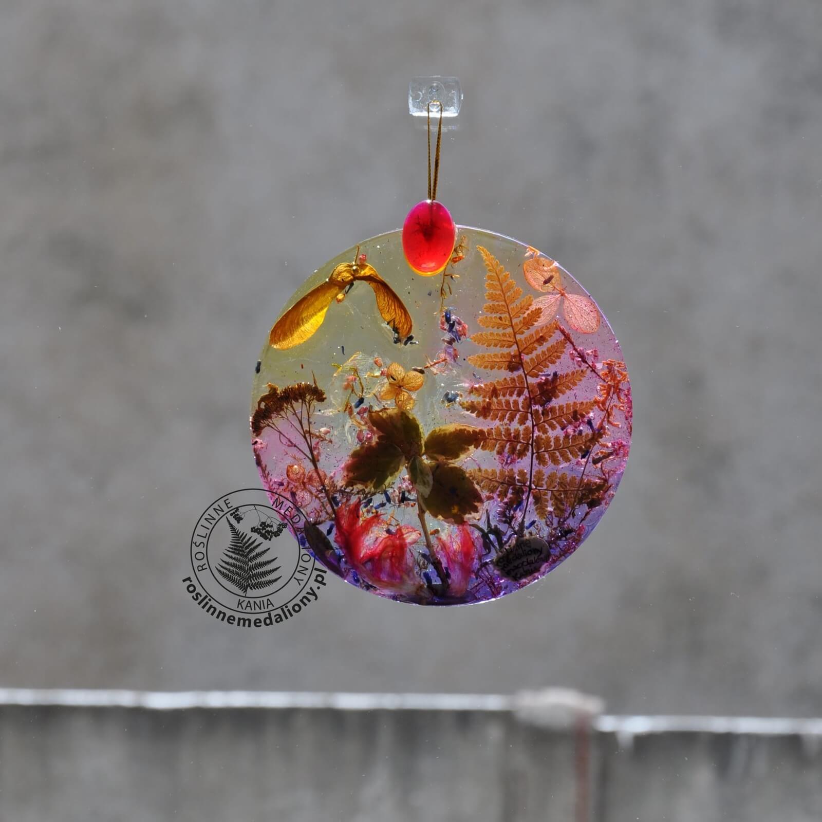 Ręcznie wykonany, okrągły medalion z żywicy, w którym zatopiono różnobarwne suszone rośliny, w tym liście paproci i bratki, zawieszony na szybie okna.