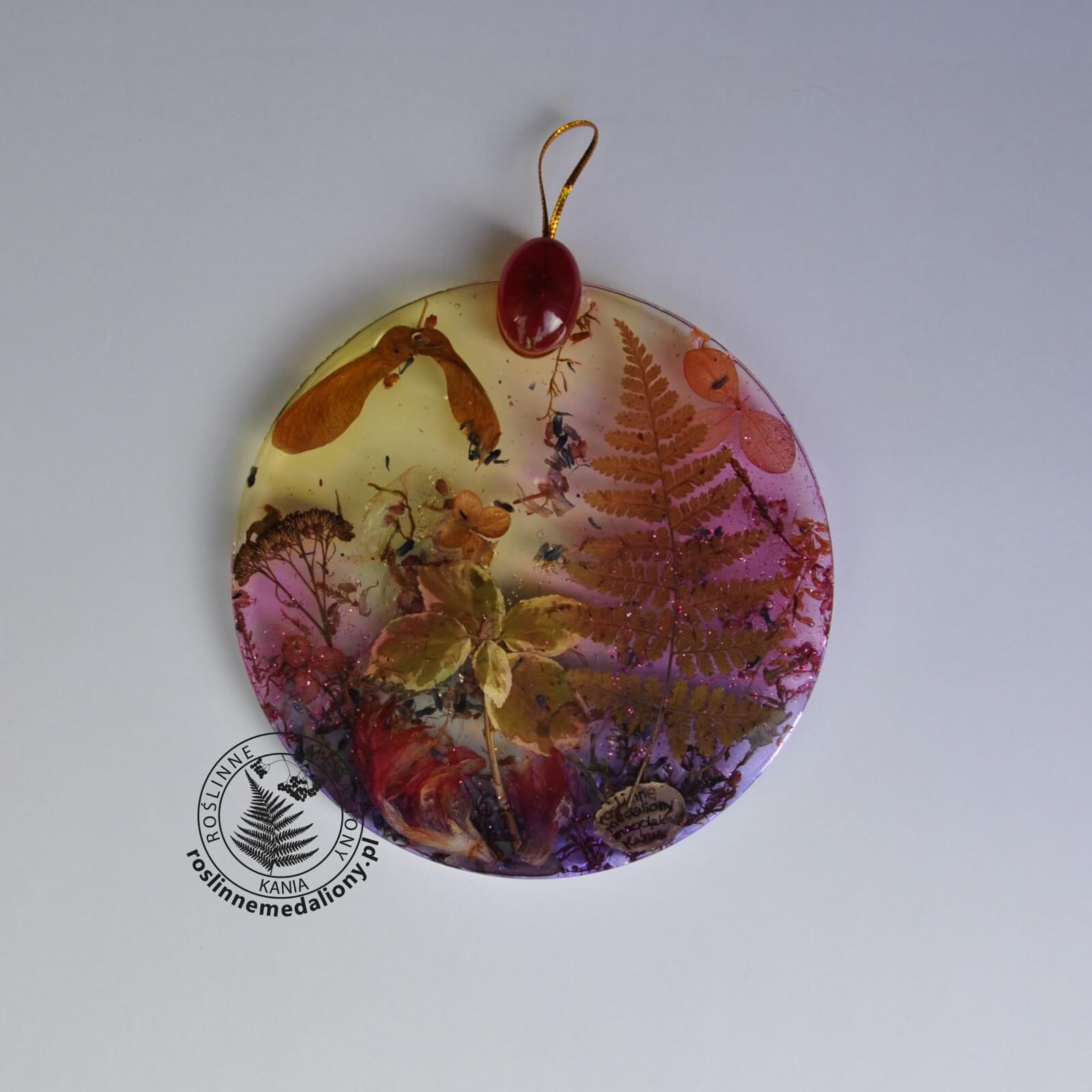 Ręcznie wykonany, okrągły medalion z żywicy, w którym zatopiono różnobarwne suszone rośliny, umieszczony na kartce papieru.