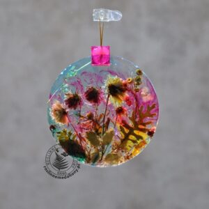 Ręcznie wykonany, okrągły medalion z żywicy, w którym zatopiono różnobarwne suszone rośliny, w tym liście paproci i bratki, zawieszony na szybie okna.