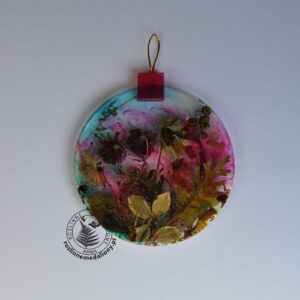 Ręcznie wykonany, okrągły medalion z żywicy, w którym zatopiono różnobarwne suszone rośliny, umieszczony na kartce papieru.