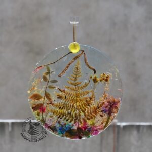 Ręcznie wykonany, okrągły medalion z żywicy, w którym zatopiono różnobarwne suszone rośliny, w tym liście paproci i bratki, zawieszony na szybie okna.