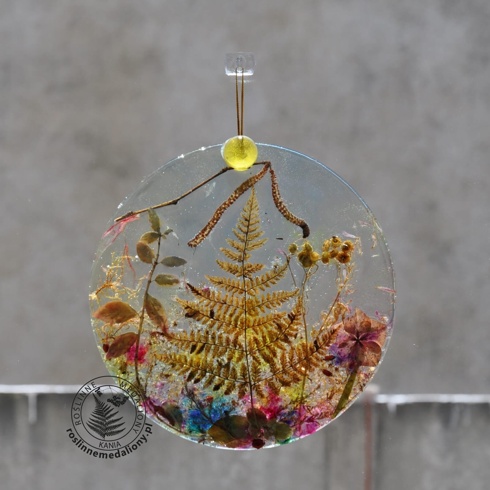 Ręcznie wykonany, okrągły medalion z żywicy, w którym zatopiono różnobarwne suszone rośliny, w tym liście paproci i bratki, zawieszony na szybie okna.