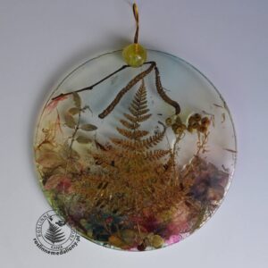Ręcznie wykonany, okrągły medalion z żywicy, w którym zatopiono różnobarwne suszone rośliny, umieszczony na kartce papieru.