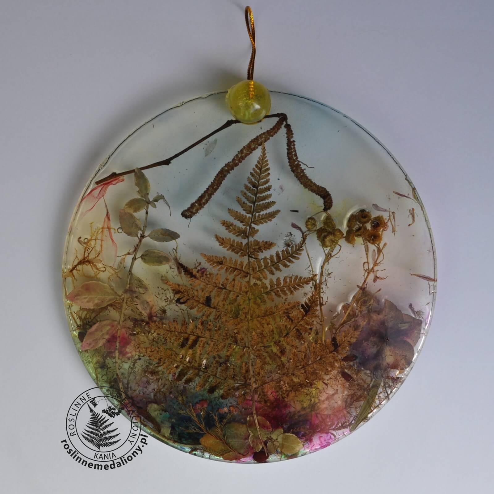 Ręcznie wykonany, okrągły medalion z żywicy, w którym zatopiono różnobarwne suszone rośliny, umieszczony na kartce papieru.