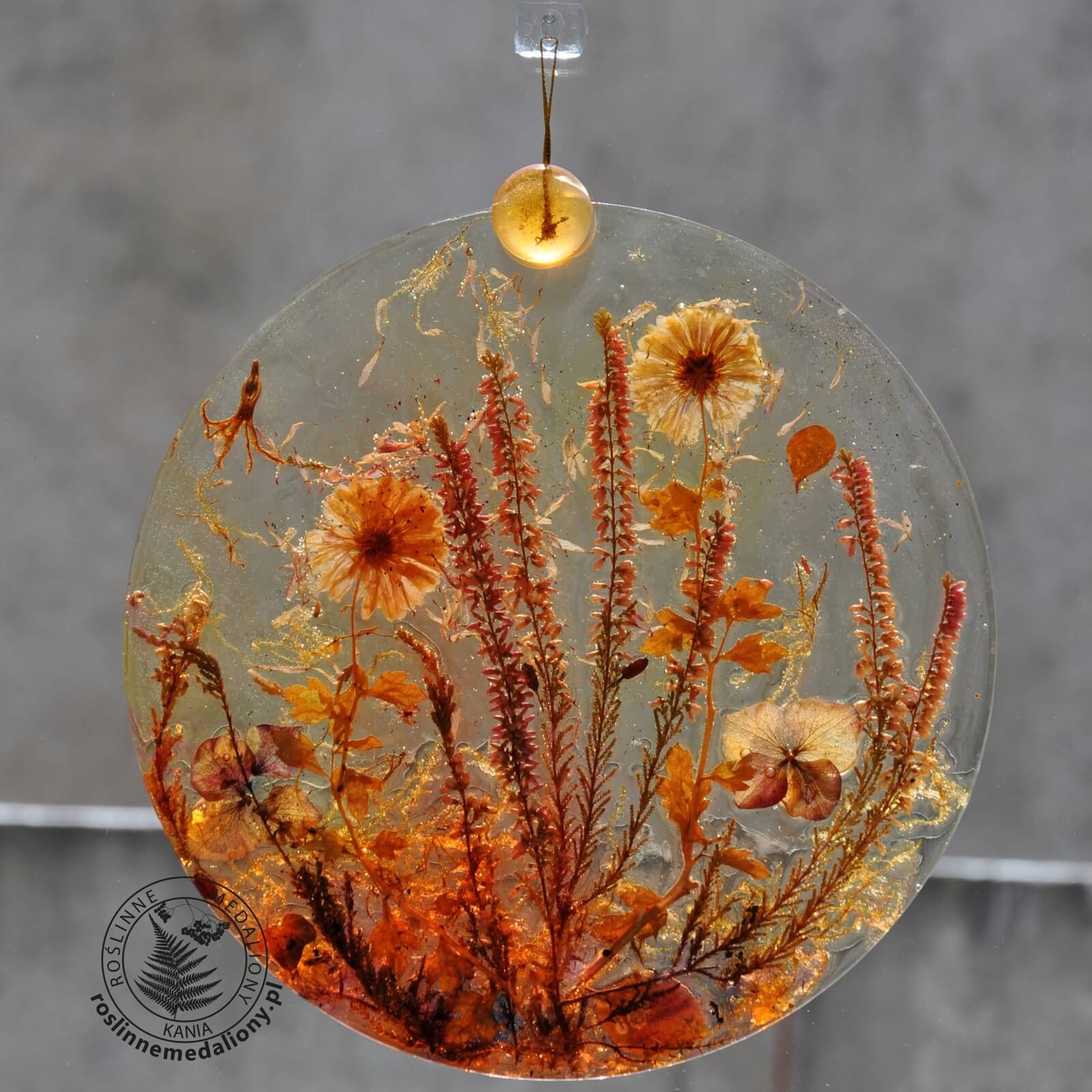 Ręcznie wykonany, okrągły medalion z żywicy, w którym zatopiono różnobarwne suszone rośliny, w tym liście paproci i bratki, zawieszony na szybie okna.