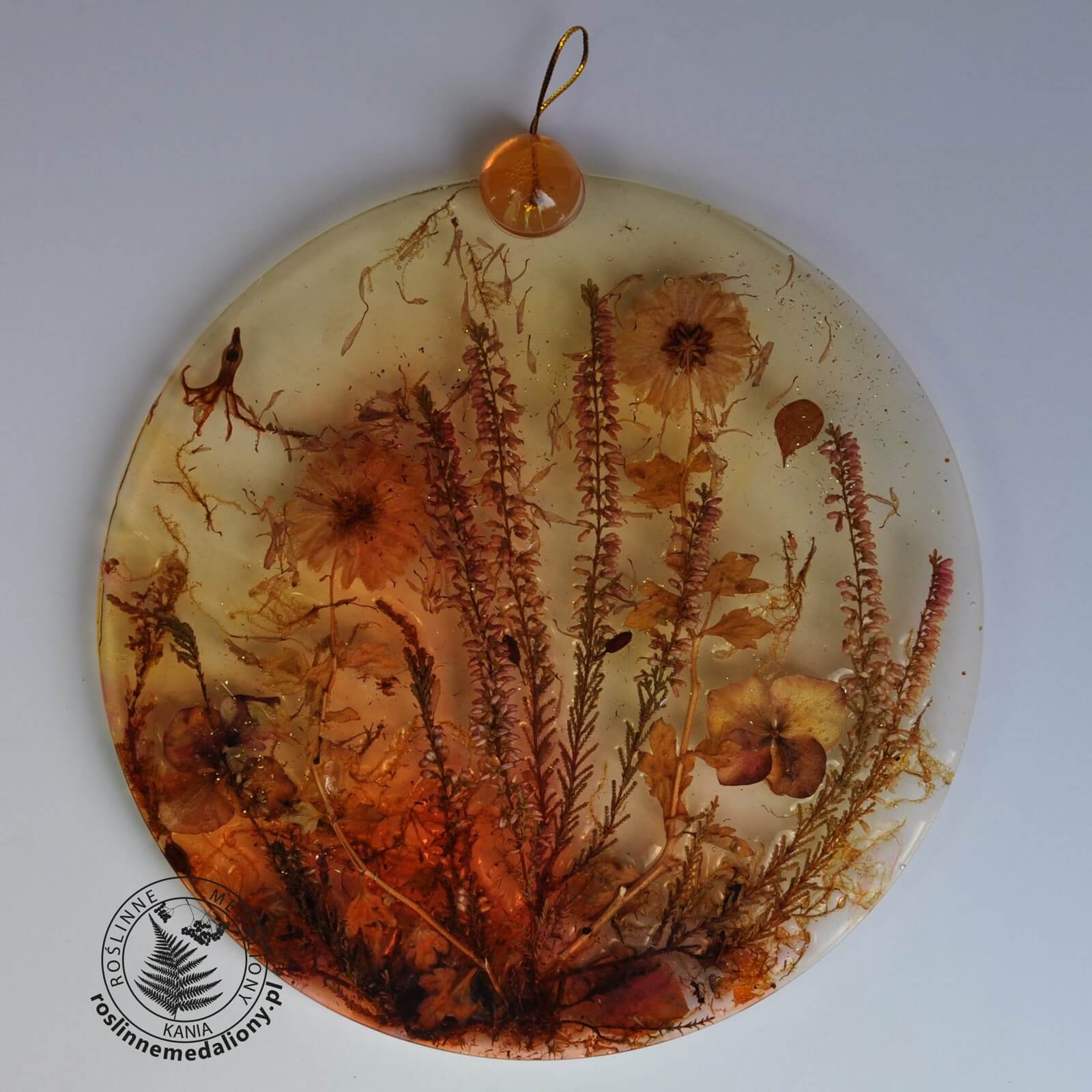 Ręcznie wykonany, okrągły medalion z żywicy, w którym zatopiono różnobarwne suszone rośliny, umieszczony na kartce papieru.