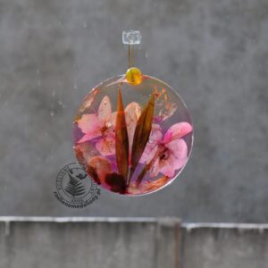 Ręcznie wykonany, okrągły medalion z żywicy, w którym zatopiono różnobarwne suszone rośliny, w tym liście paproci i bratki, zawieszony na szybie okna.