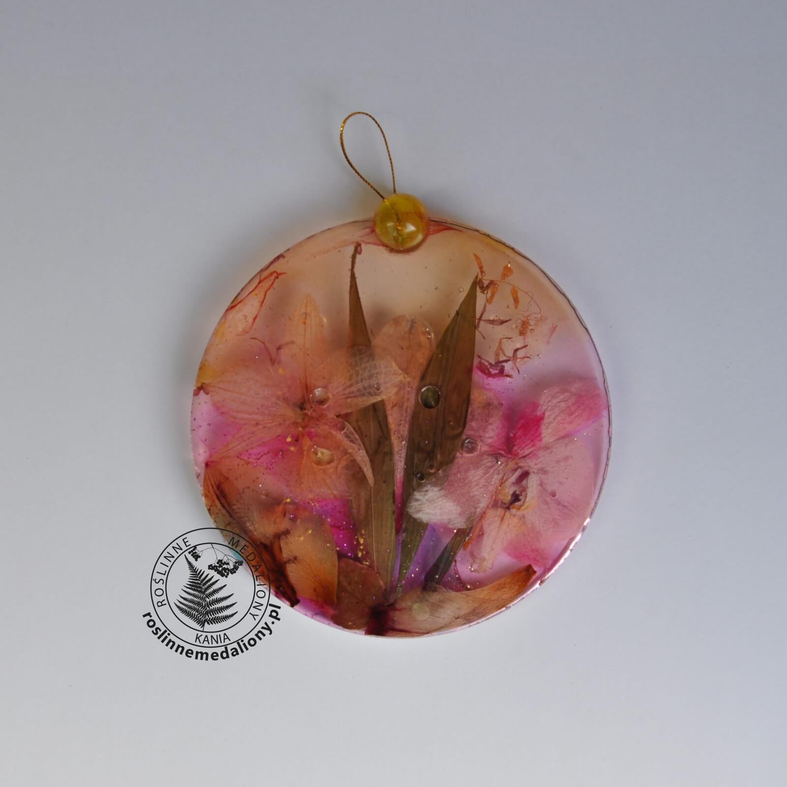 Ręcznie wykonany, okrągły medalion z żywicy, w którym zatopiono różnobarwne suszone rośliny, umieszczony na kartce papieru.